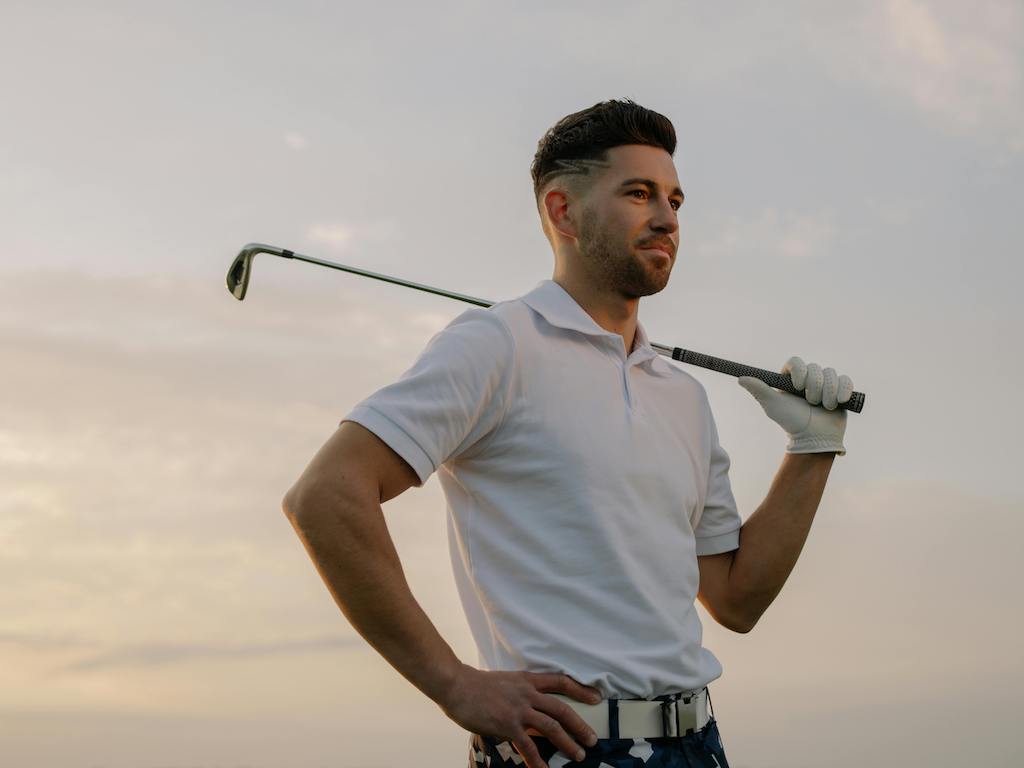 man holding a golf club wearing a polo, belt, and pants